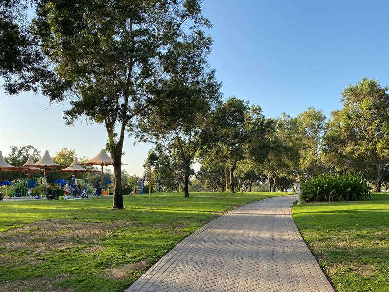 Grüner Stadtpark. Es gibt ein Weg um durchzulaufen. Am Wegrand stehen mehrere Bäume. Links im Hintergrund stehen Sonnenschirme.