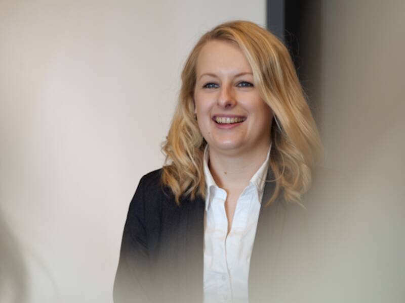 Portrait einer Frau mit schulterlangen, blonden und welligen Haaren. Sie trägt eine weiße Bluse und darüber einen schwarzen Blazer.